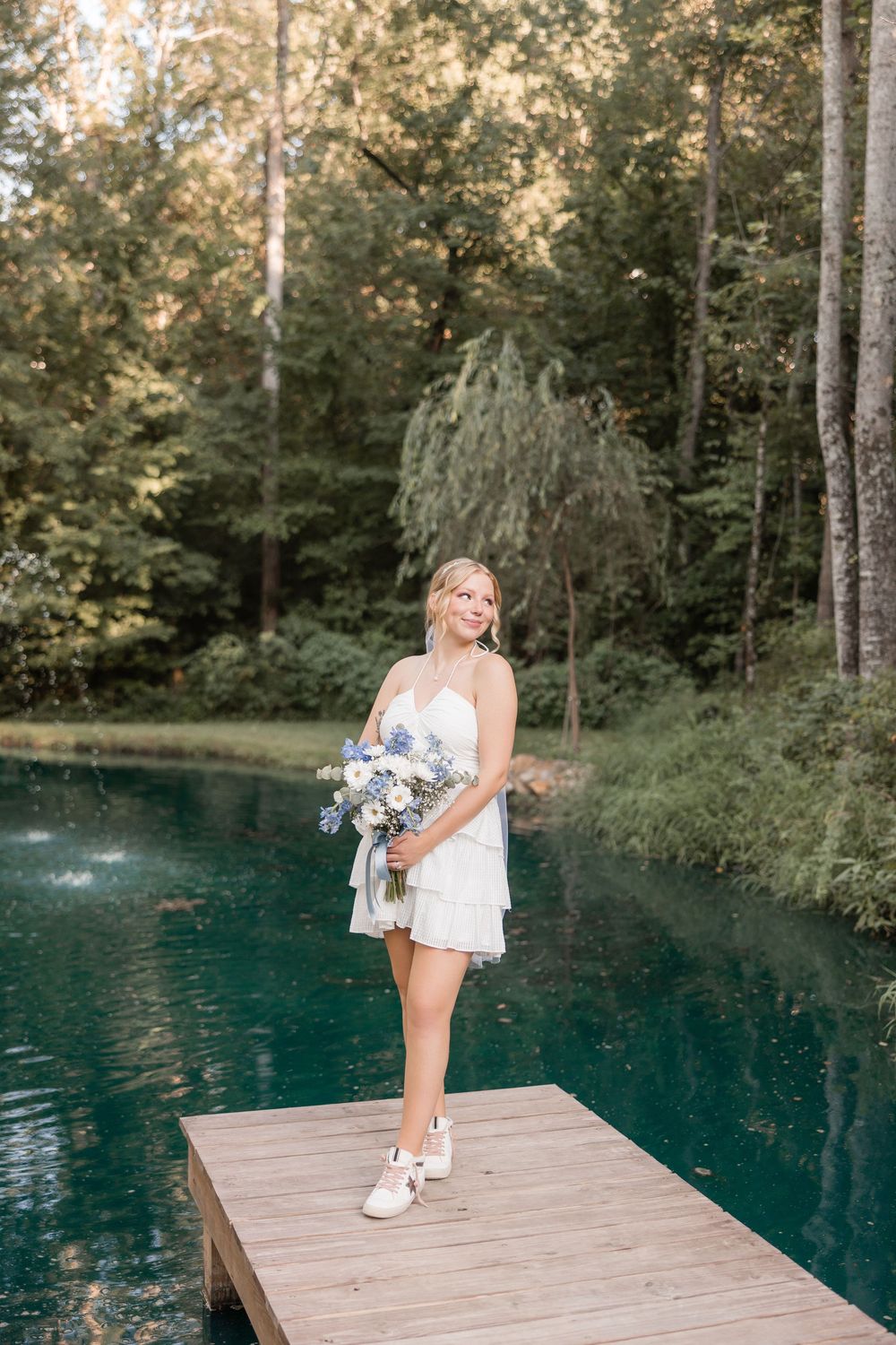 Woman in a white dress poses on a wooden dock overlooking a serene blue spring pond surrounded by forest.