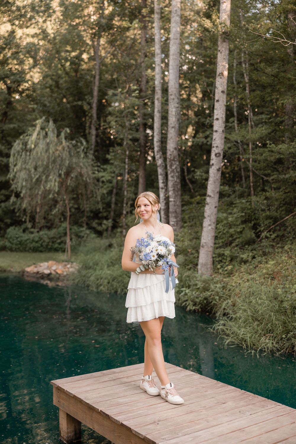 A person in a summer dress sits on a wooden deck surrounded by trees and reflective blue water.
