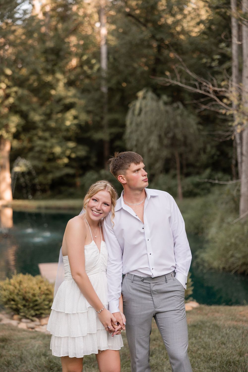 A couple poses playfully in a grassy outdoor setting near a pond.