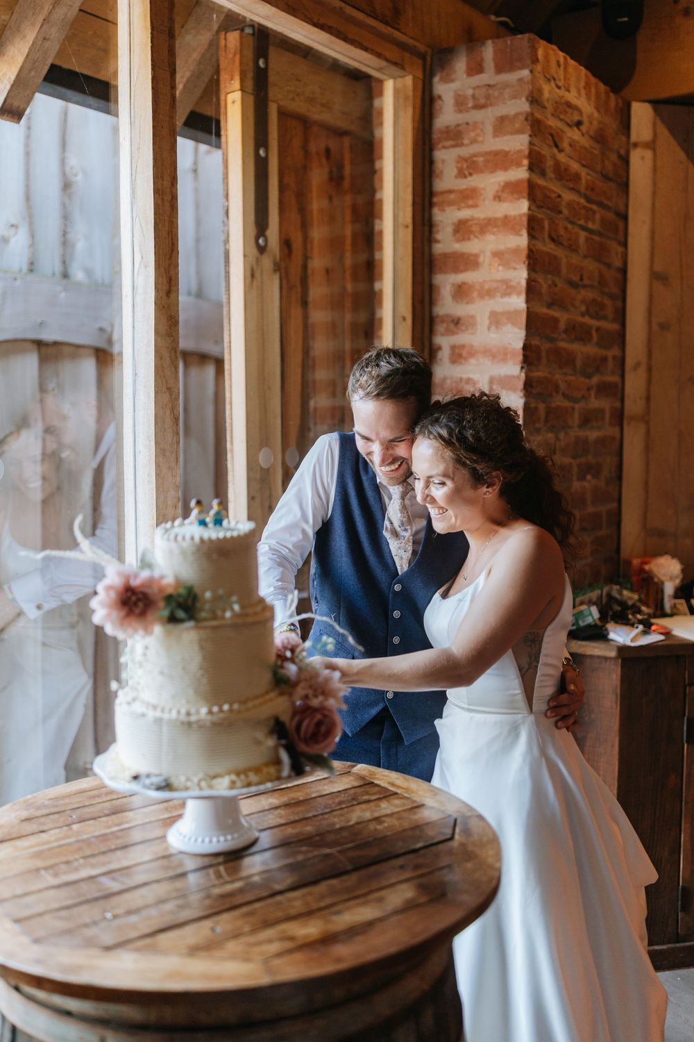 Bride and groom smiling while cutting their three-tier white wedding cake in a rustic barn venue.