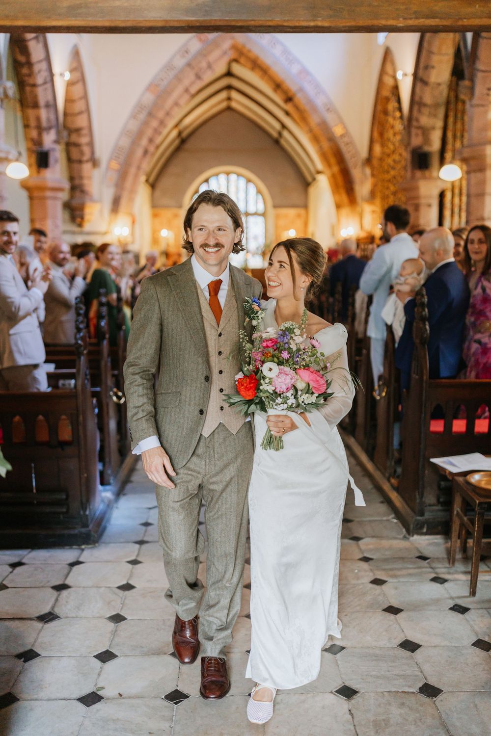 Newlywed couple walking down church aisle, bride holding colorful bouquet in white gown, groom in tweed suit with orange tie.