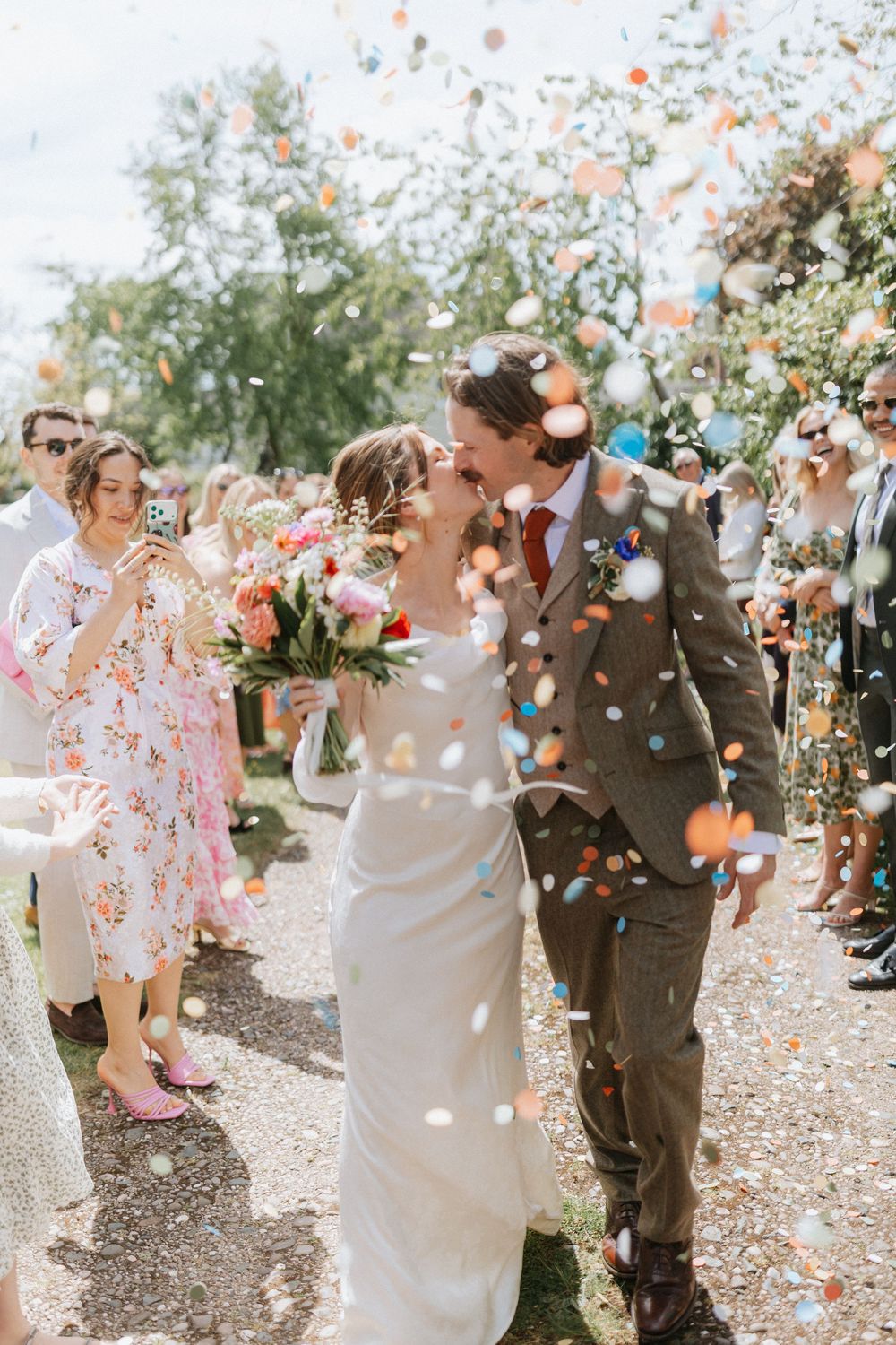 Bride and groom share a kiss while colorful confetti fills the air during their outdoor wedding ceremony.