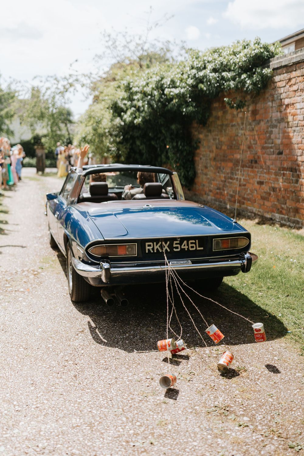 Blue vintage Triumph convertible wedding car with tin cans tied to bumper on gravel driveway.