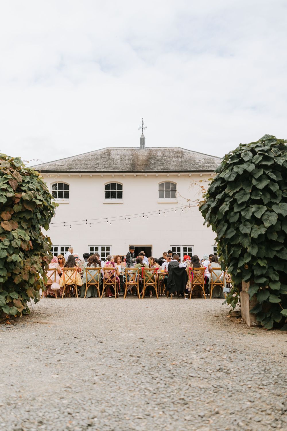 Long dining tables and chairs set up outdoors against a white building wall with string lights and climbing vines.