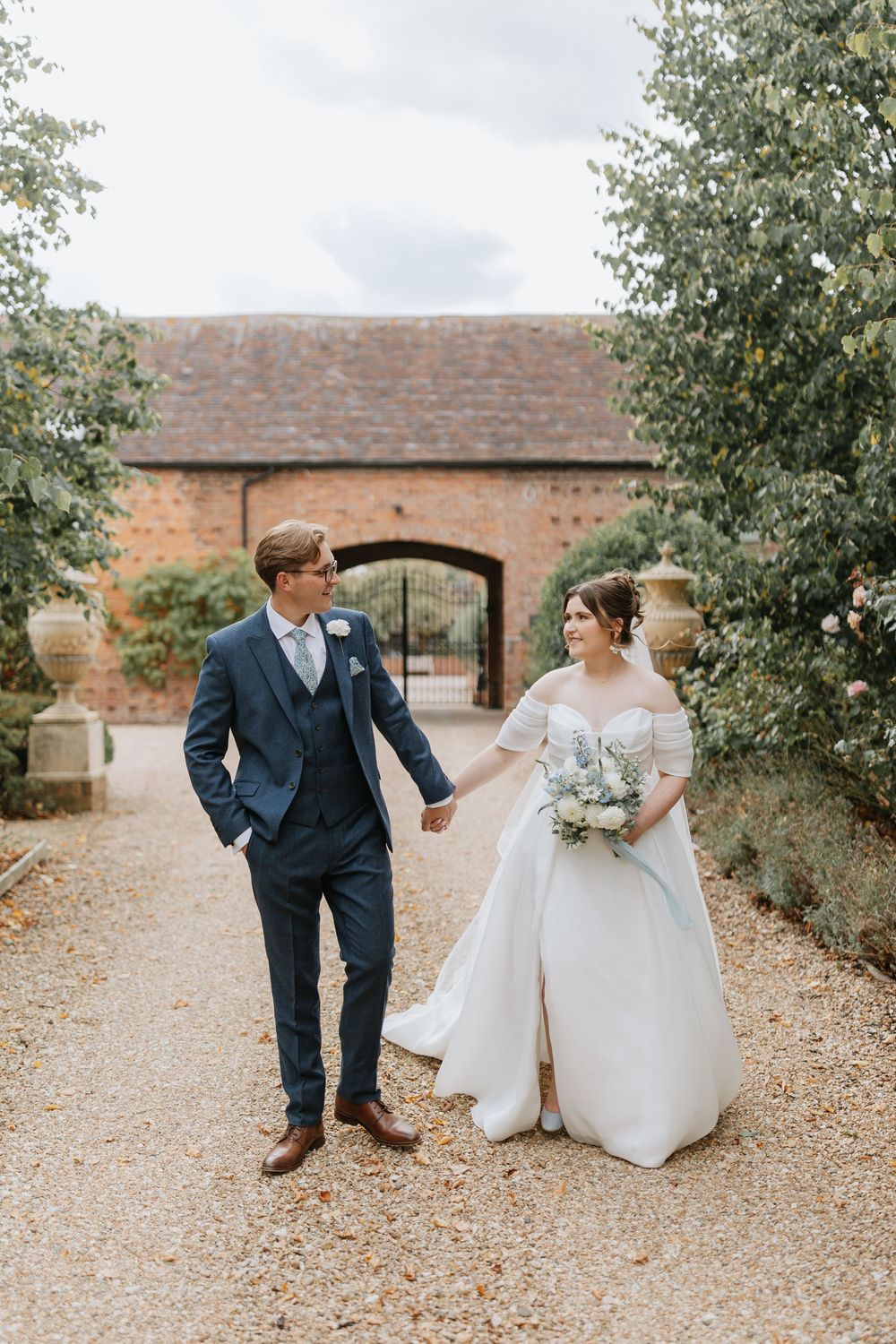 Bride and groom hold hands walking on gravel path at elegant brick venue, bride in off-shoulder gown with bouquet. Hencote, Shropshire