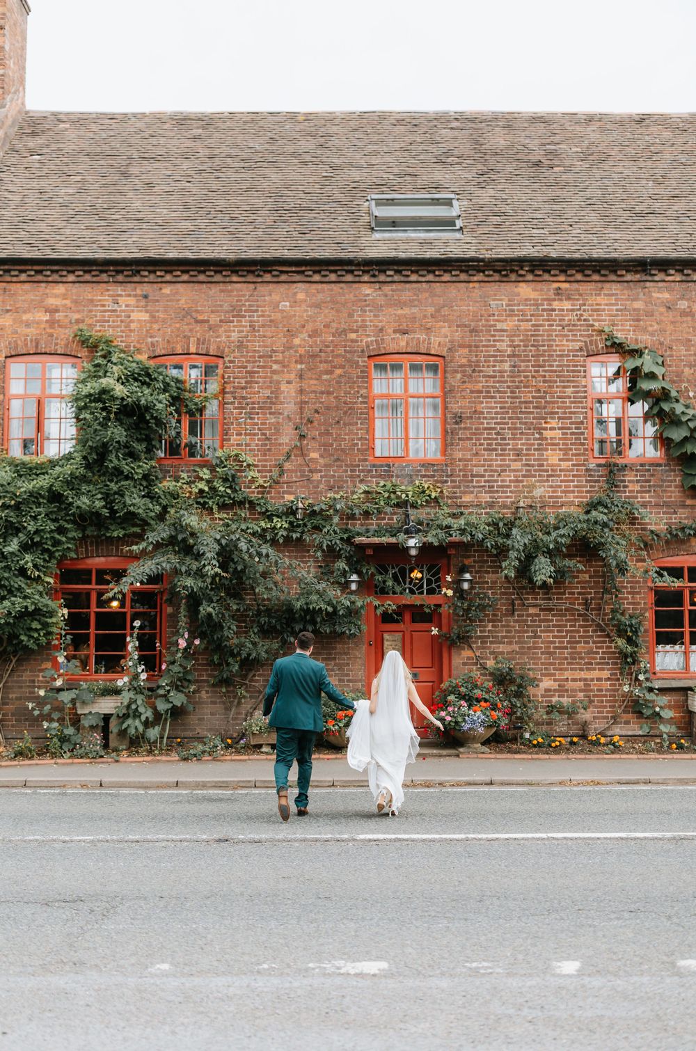 Bride and groom walking hand in hand toward a charming ivy-covered red brick building on their wedding day.
