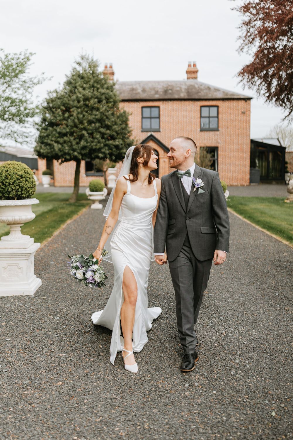 Bride and groom walking hand in hand outside a brick manor house, bride holding a bouquet in a white gown.