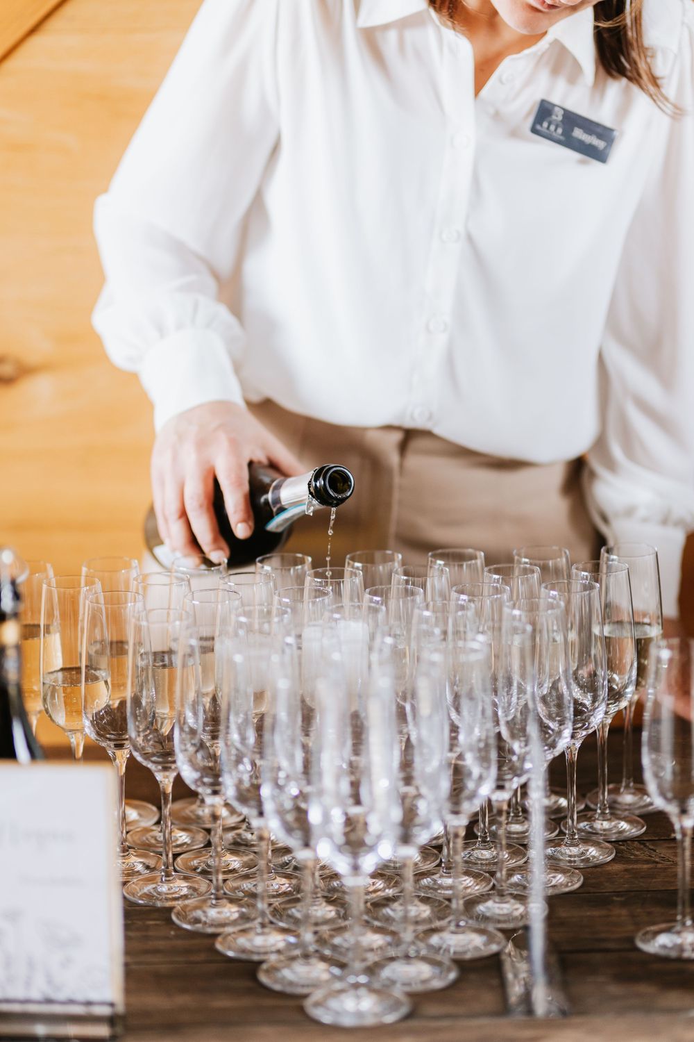 A waitress in a white shirt pours champagne into rows of elegant crystal flute glasses at a formal event.