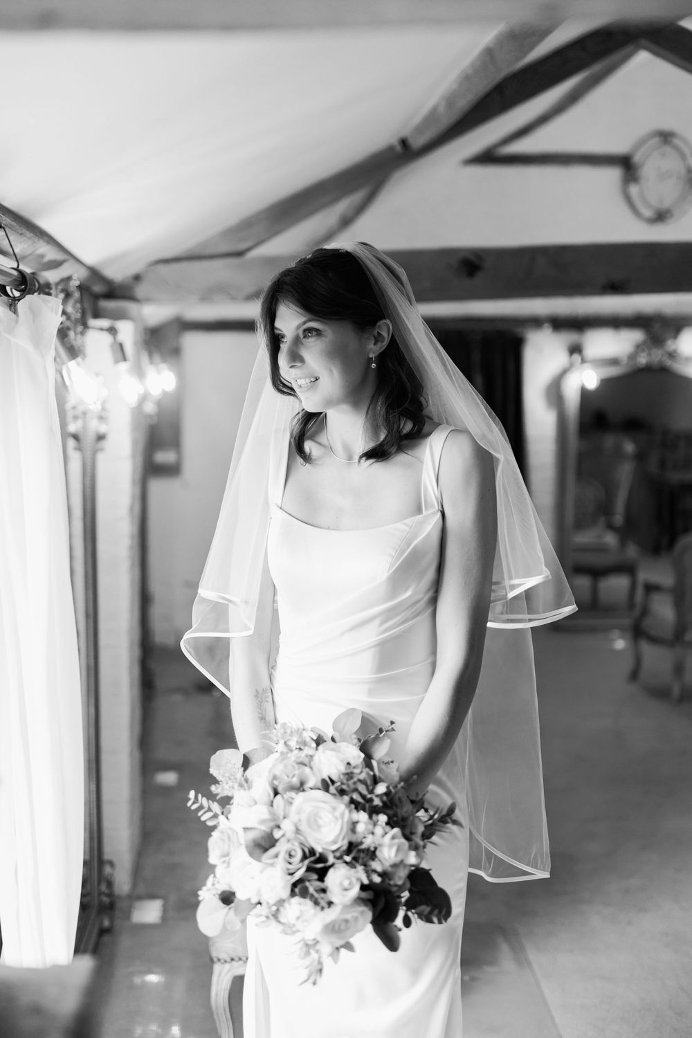 Black and white photo of a bride in a sleek gown and veil holding a bouquet of roses indoors.