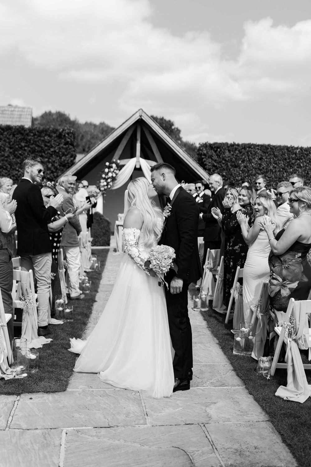 Black and white photo of bride and groom kissing at outdoor wedding ceremony aisle with guests applauding.