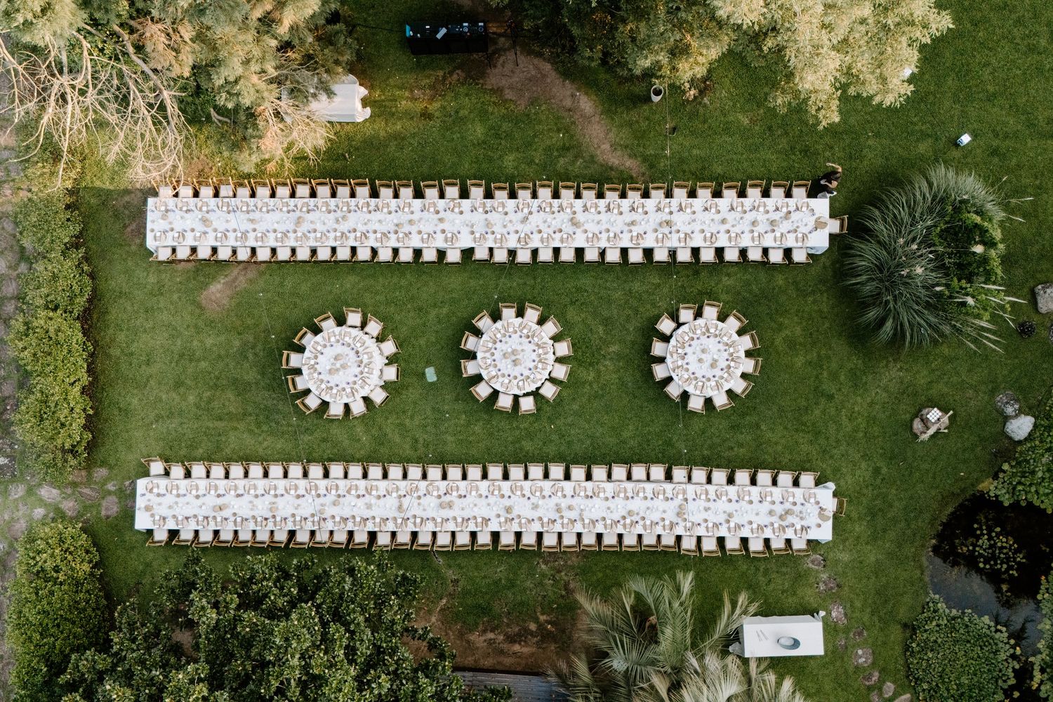 Vista aérea de elegantes mesas blancas de recepción de bodas dispuestas en filas sobre un césped verde. Jardins el Roquer