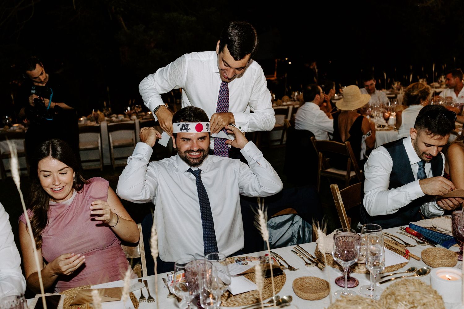 Wedding guests enjoy a lively dinner reception under warm lighting at elegantly set tables.