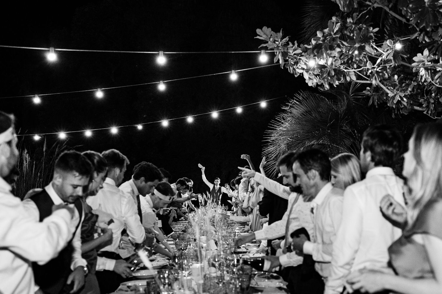 String lights glow above a lively outdoor wedding reception celebration.