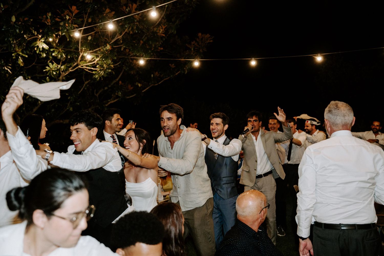 Wedding guests dancing joyfully under string lights at an outdoor nighttime celebration.
