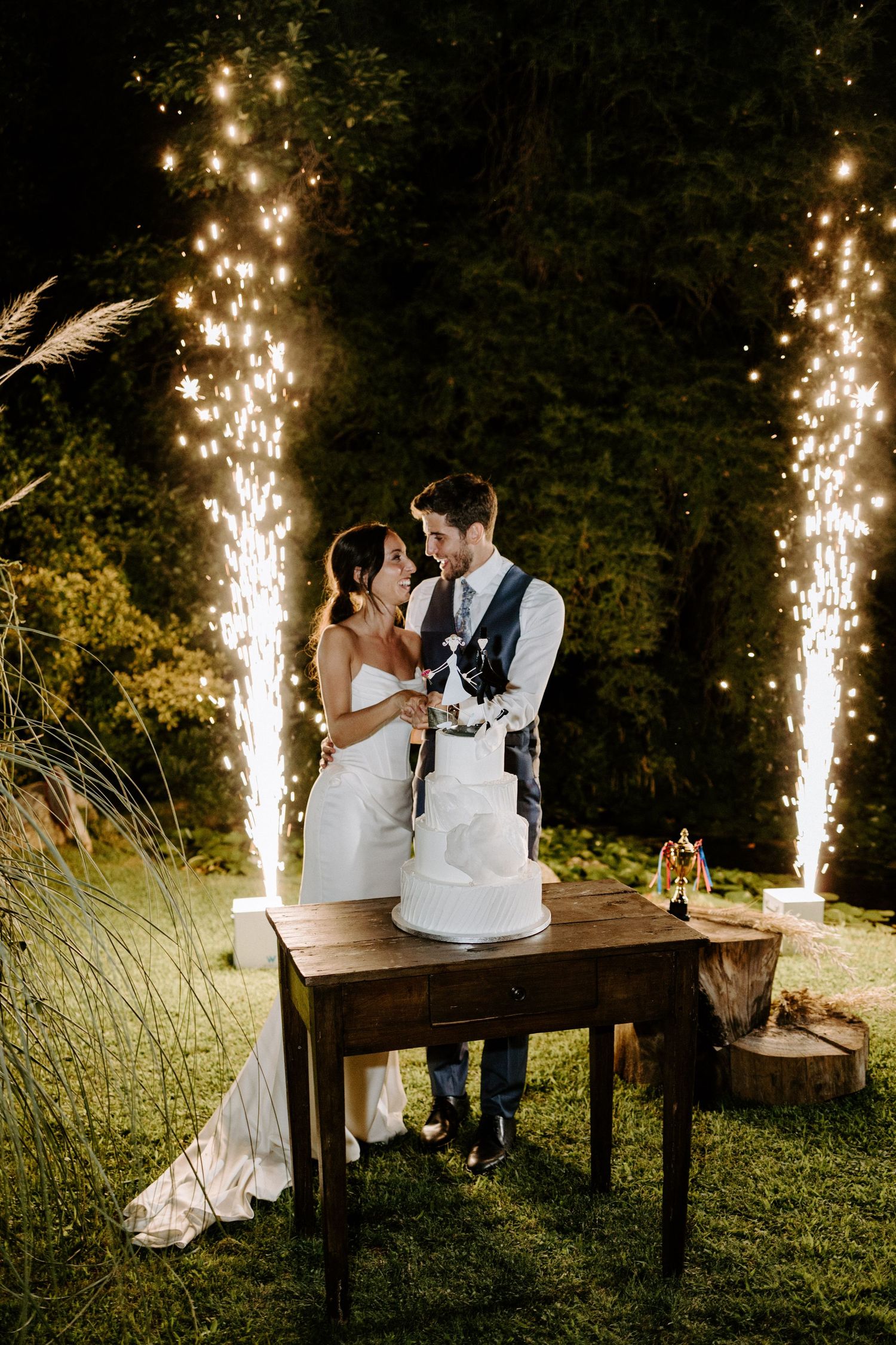 Couple cutting wedding cake on rustic table with dramatic sparkler fountains in background.