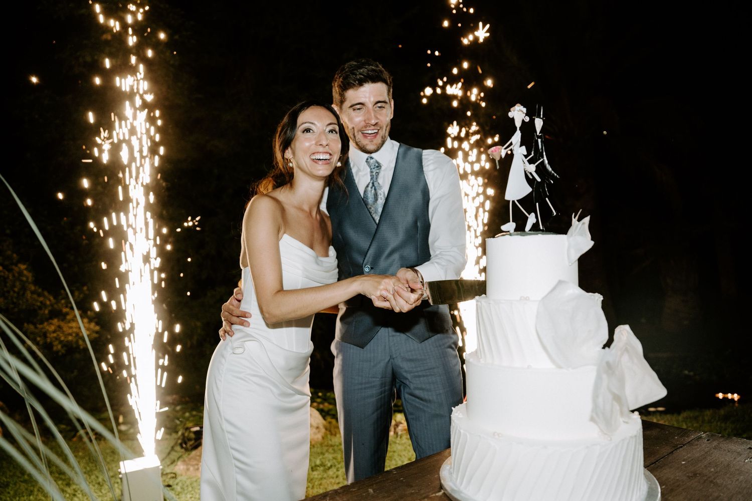 Newlyweds cutting their elegant white wedding cake with sparkler effects in the background.
