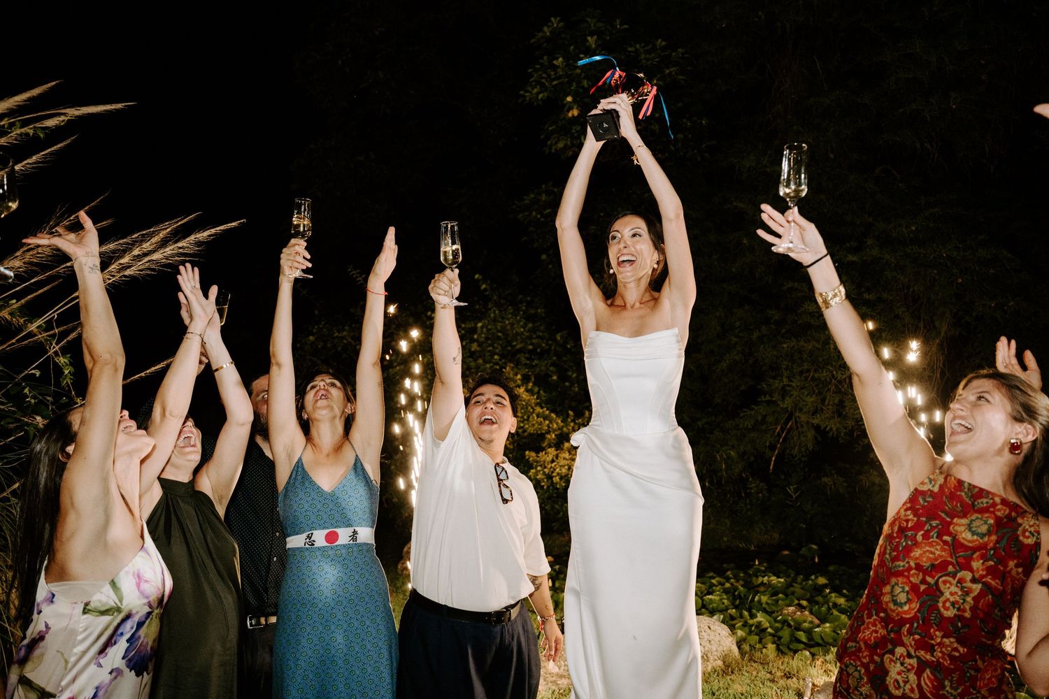 Wedding party raising champagne glasses in celebration under string lights.