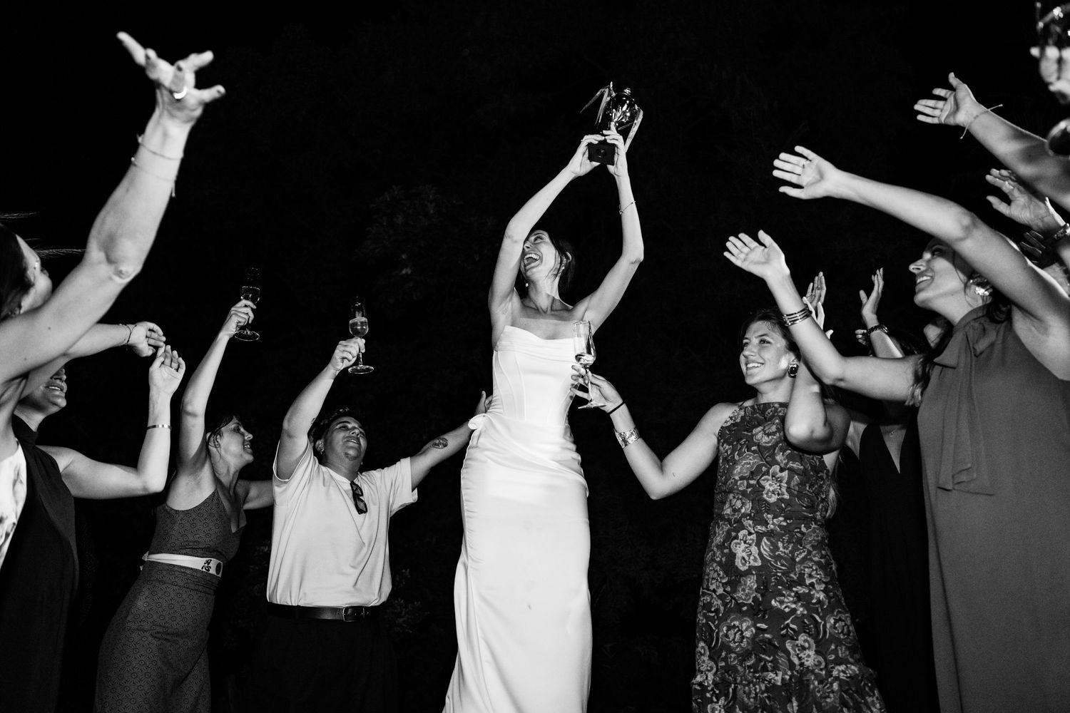 Black and white photo of wedding celebration with guests raising arms in joyful dance.