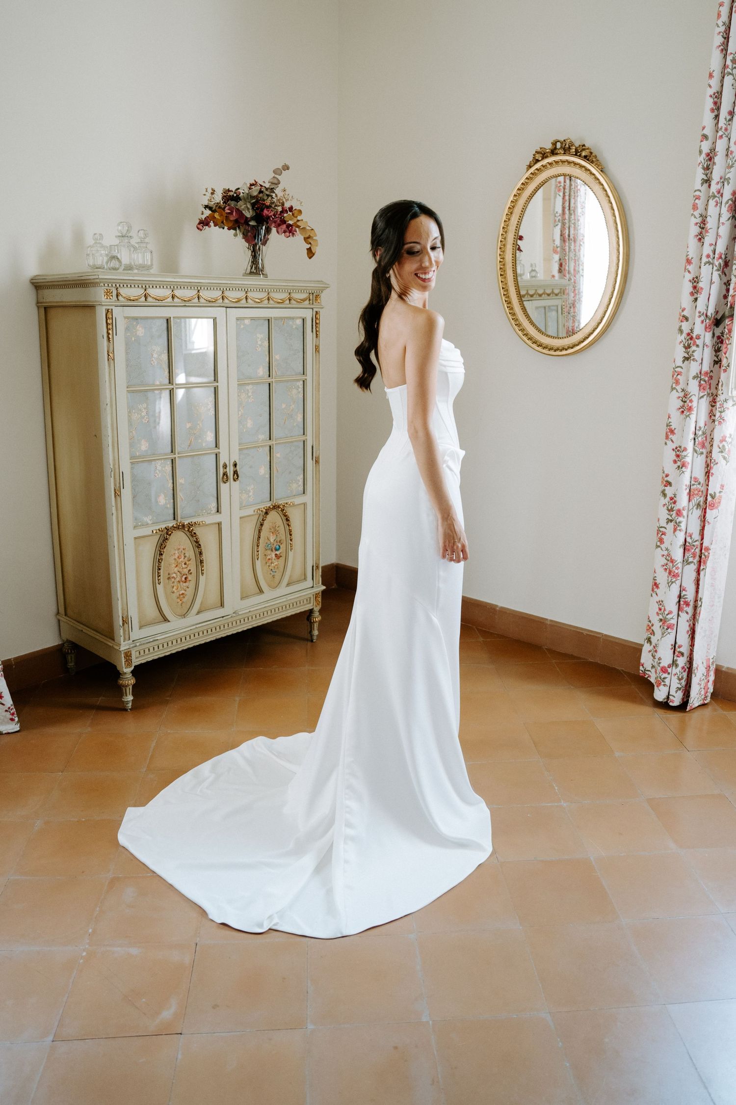 Bride in flowing white wedding gown posing in front of vintage-style furniture and floral curtains.