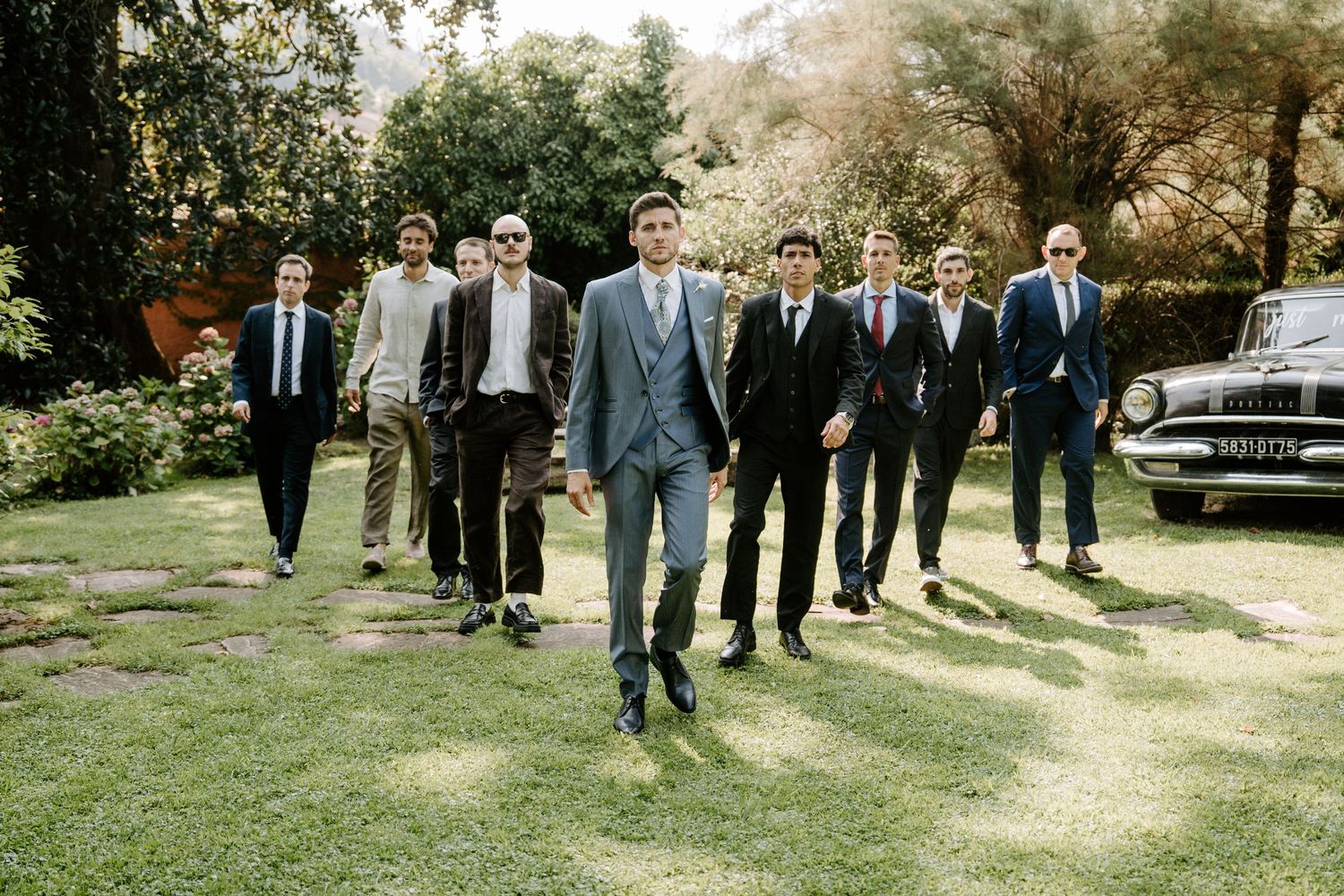 A group of groomsmen walk together on a sunlit lawn in matching dark suits and ties.