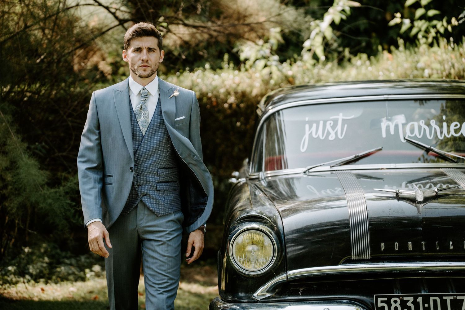 A grey suit-clad figure stands next to a vintage car with 'Just Married' written on it.