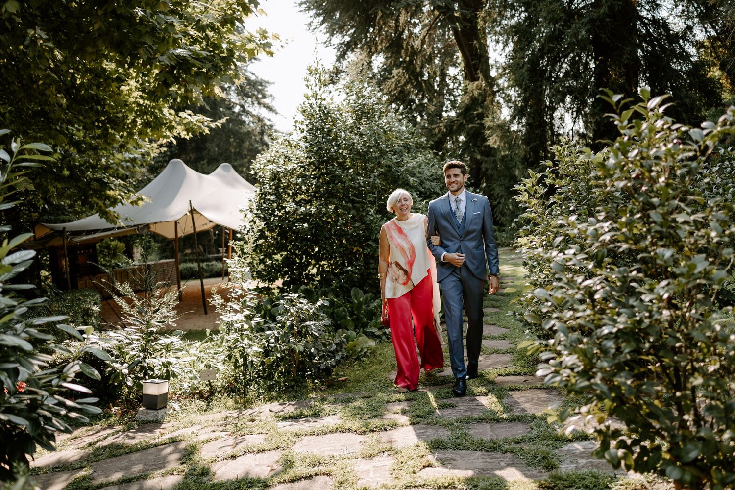 A couple walks along a garden path with white tent and greenery in background.