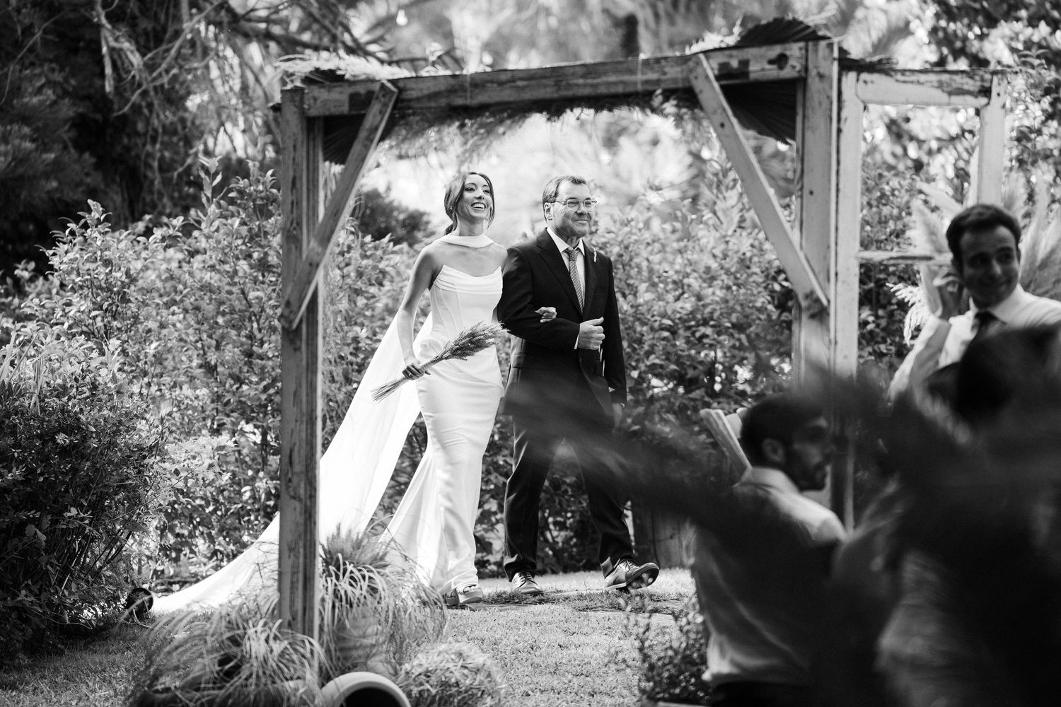 Black and white ceremony photo under a wooden arch with guests in foreground.