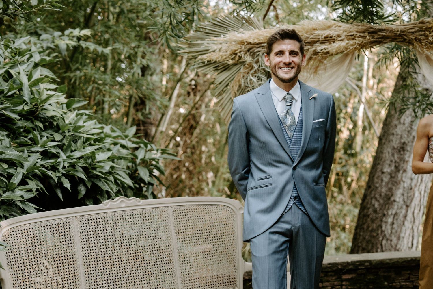 A grey suited figure stands in front of a rustic wedding arch surrounded by greenery.