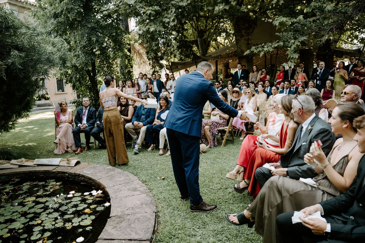 Rows of wedding guests seated outdoors under green trees during a garden ceremony.