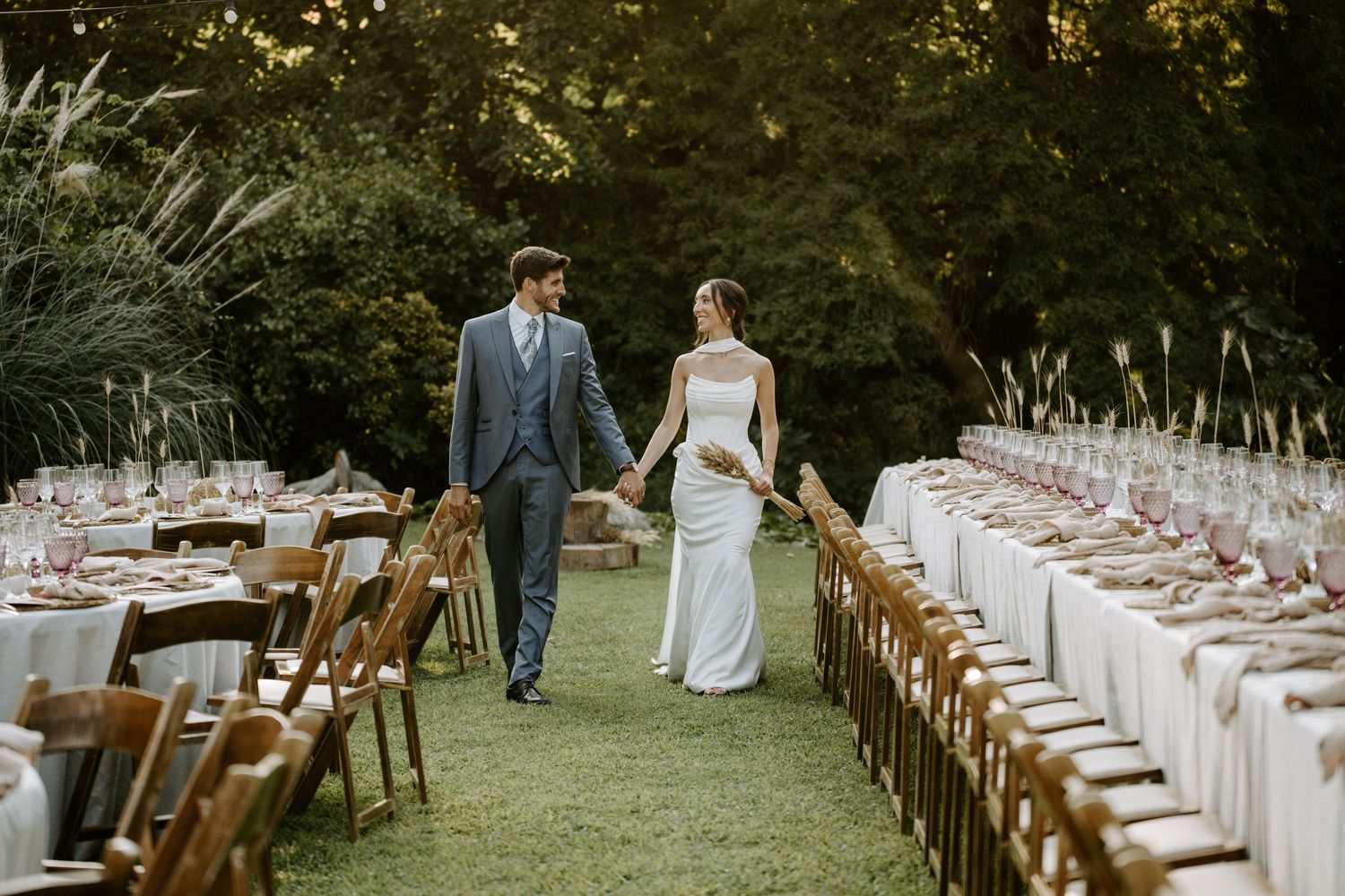 Recepción de boda al aire libre con largas mesas de madera y manteles blancos en un jardín. Jardins El Roquer