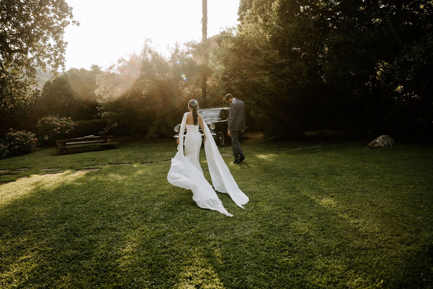 Newlyweds walk through a sunlit garden with flowing white dress trailing behind.