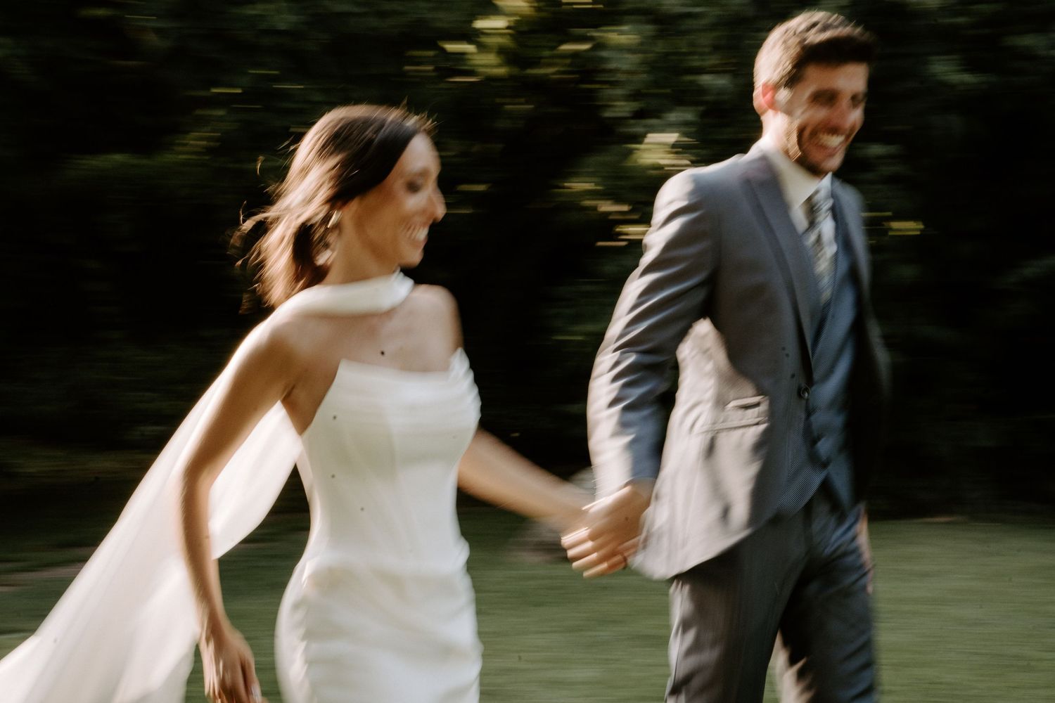 Couple in wedding attire walks together through a wooded area.