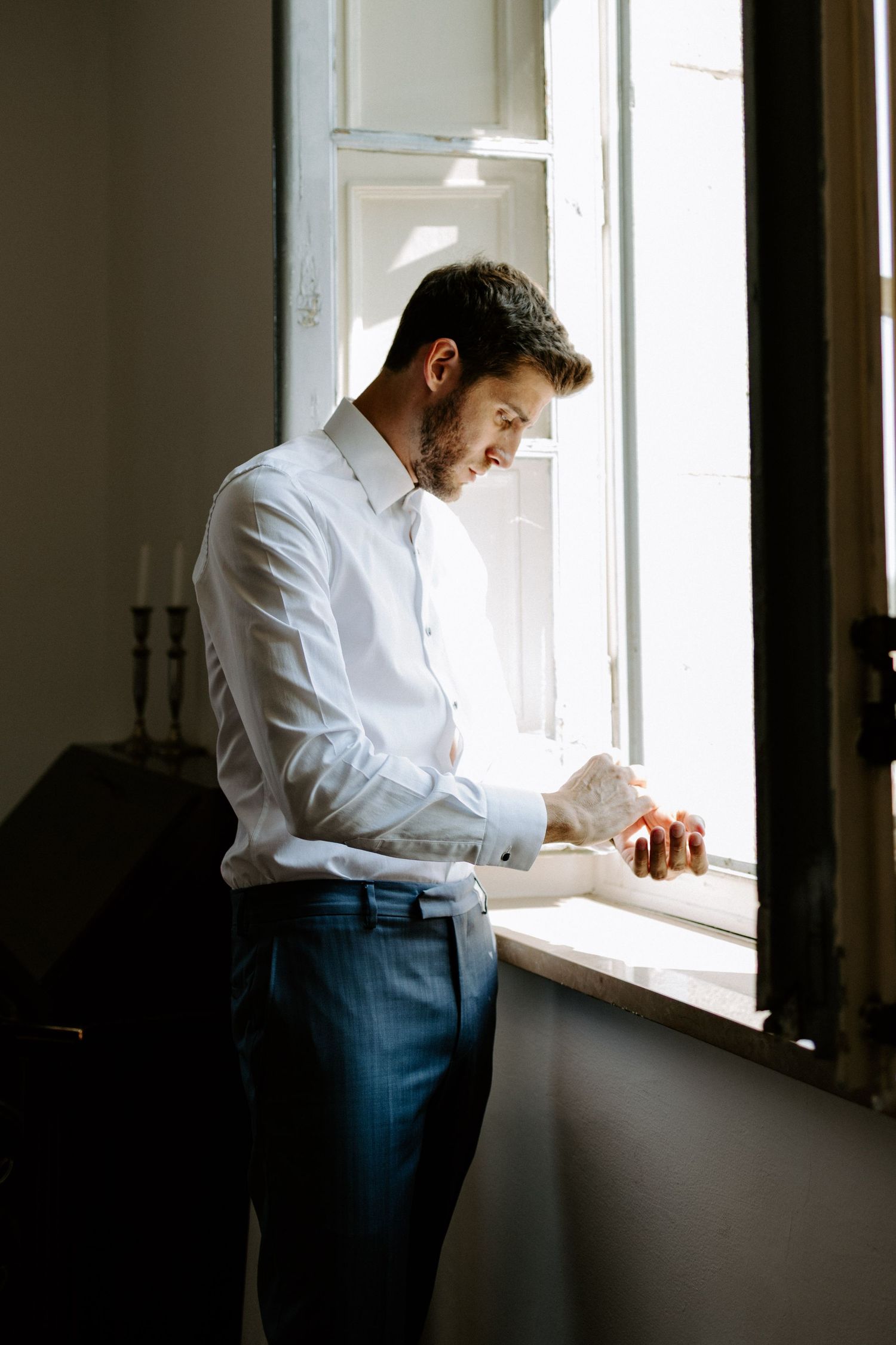 Person in white dress shirt and blue pants standing by bright window.