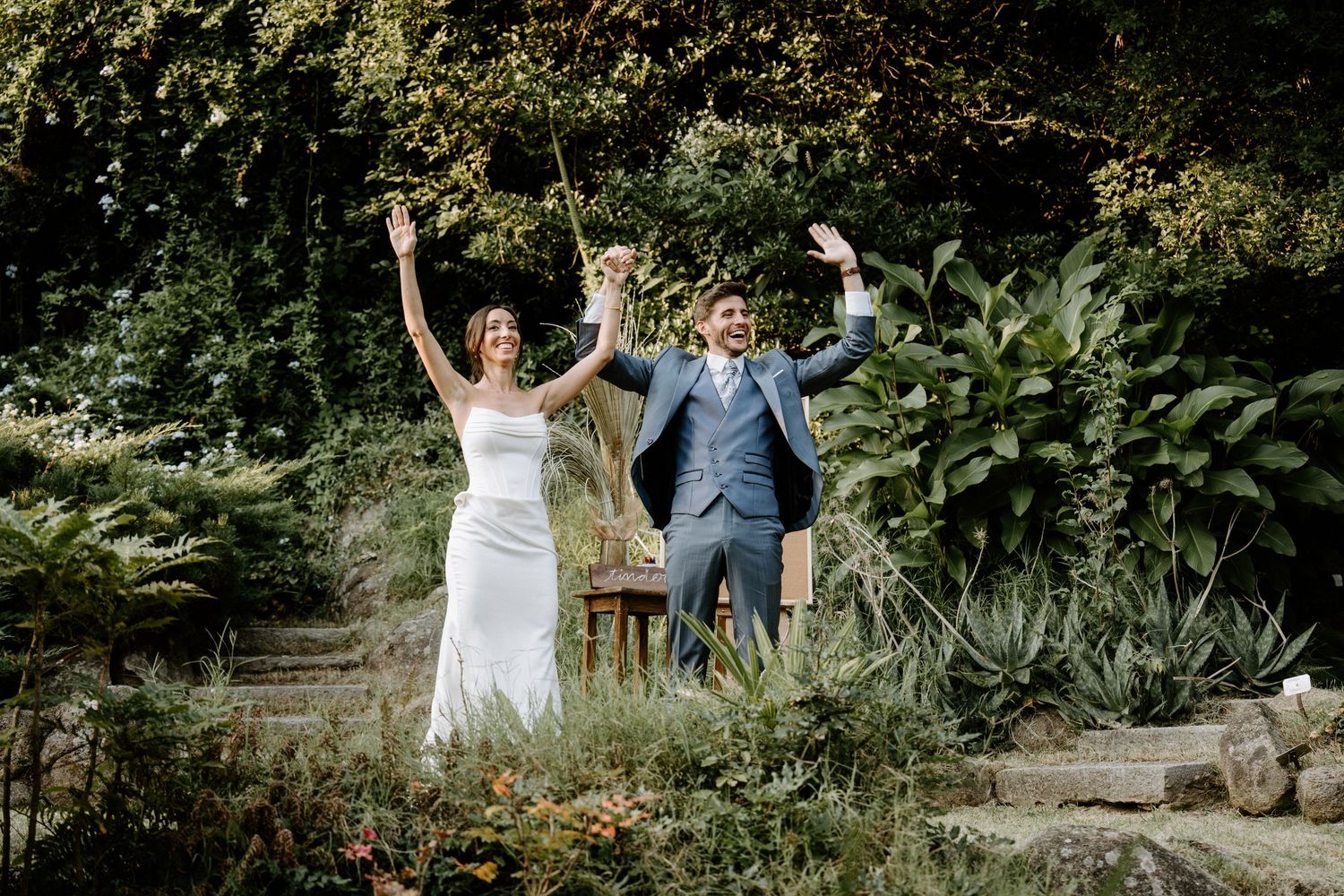 Joyful wedding couple celebrating with raised arms in a lush garden setting.
