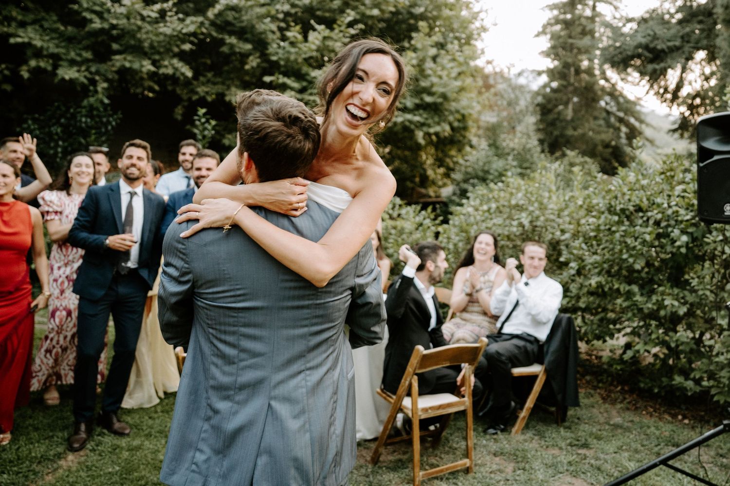 Guests celebrate and embrace during an outdoor wedding reception surrounded by trees.