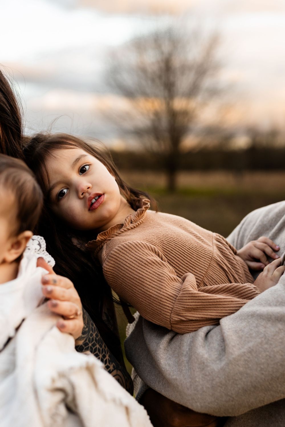 Young girl in brown ribbed top resting in mother's arms outdoors at sunset with a sibling nearby.
