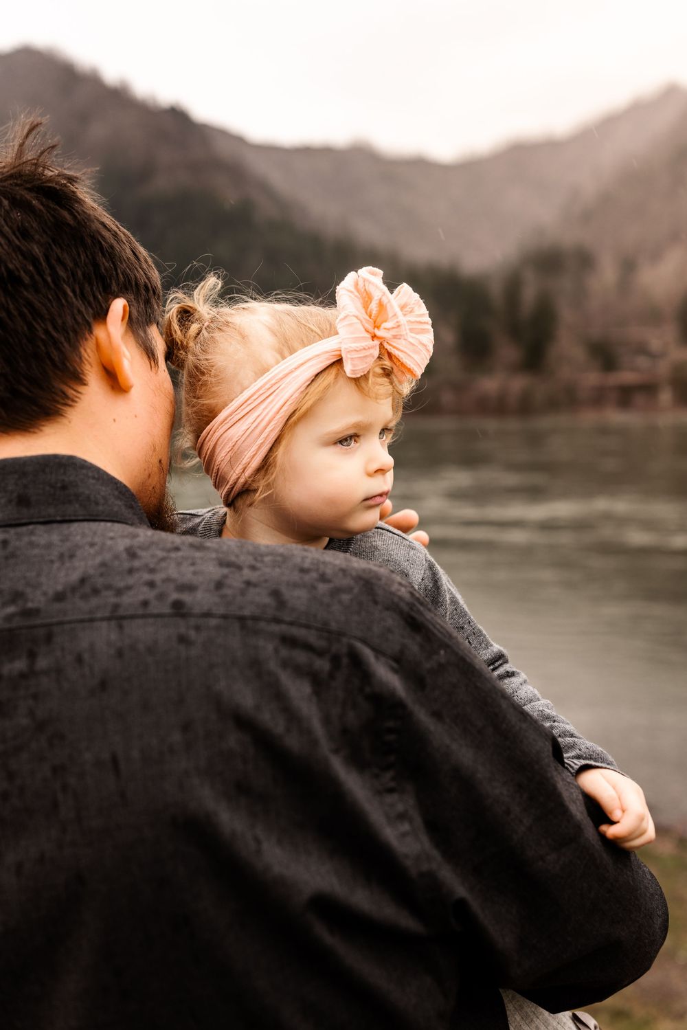 A father holds his young blonde toddler girl wearing a pink bow headband near a scenic mountain river.