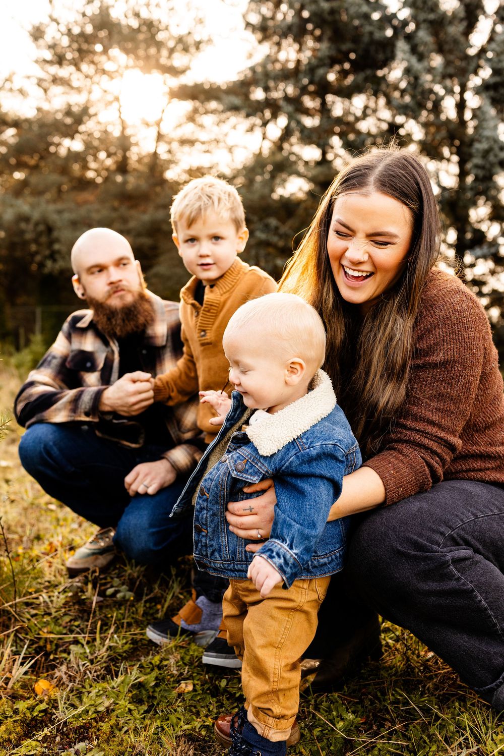 A joyful family of four poses outdoors in autumn, with parents and two young boys smiling among golden grass and trees.