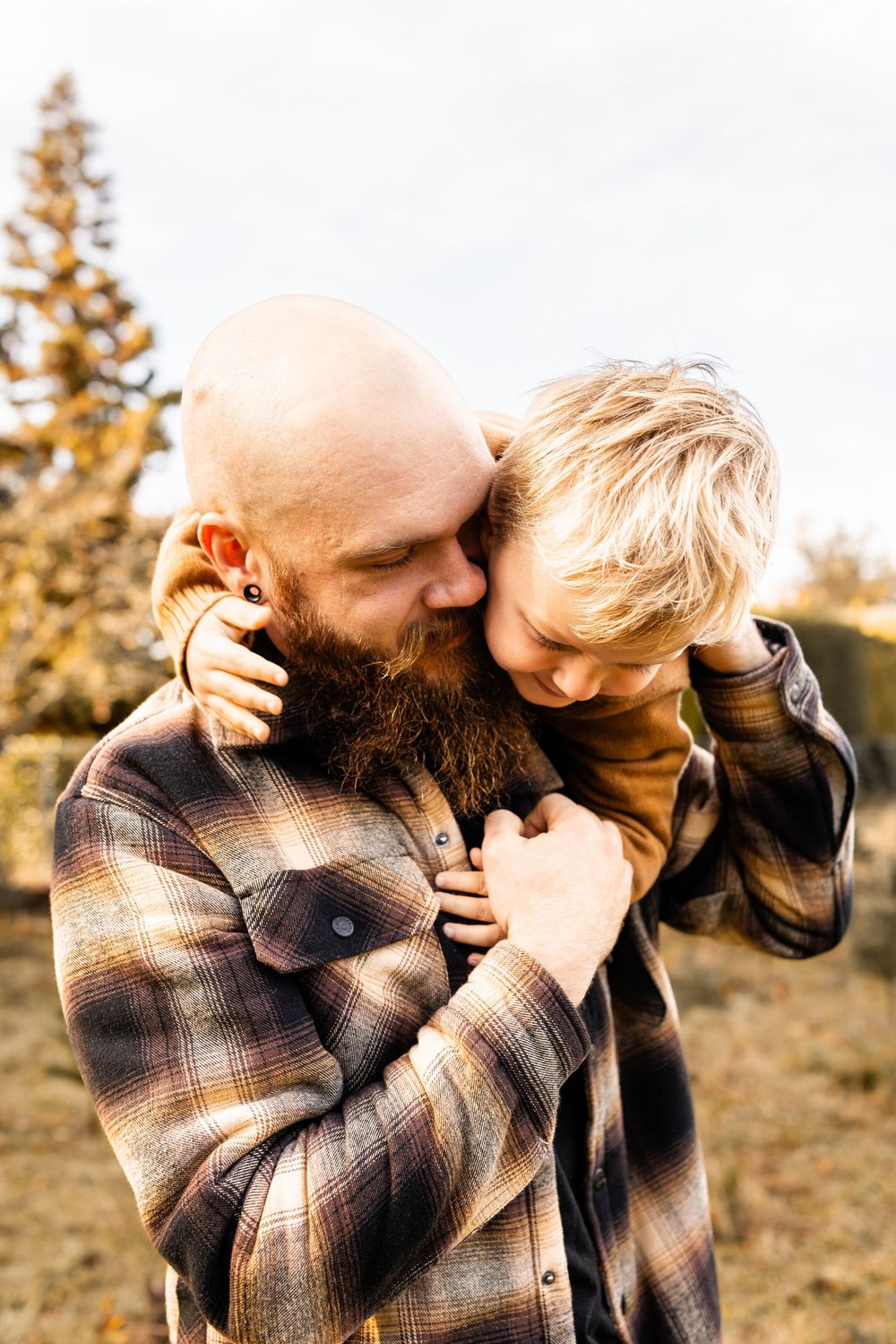 Bald bearded father in plaid shirt tenderly embracing his young blonde son outdoors in a warm autumn setting.