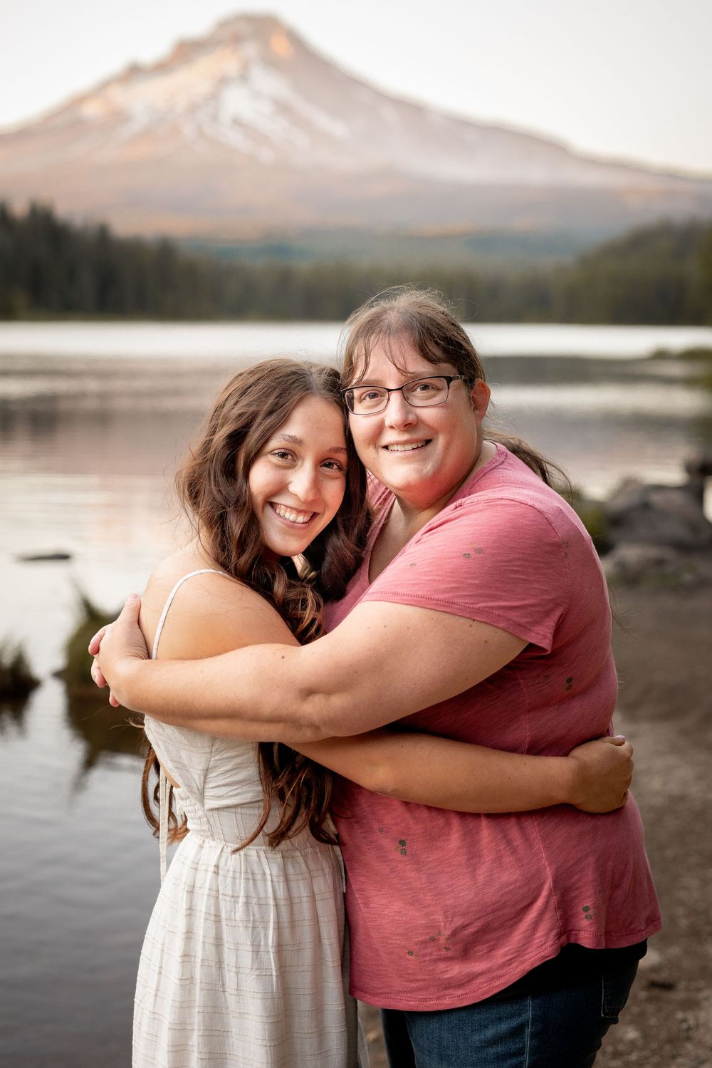 Two people embrace while smiling at Trillium Lake with Mount Hood visible in the background during sunset.