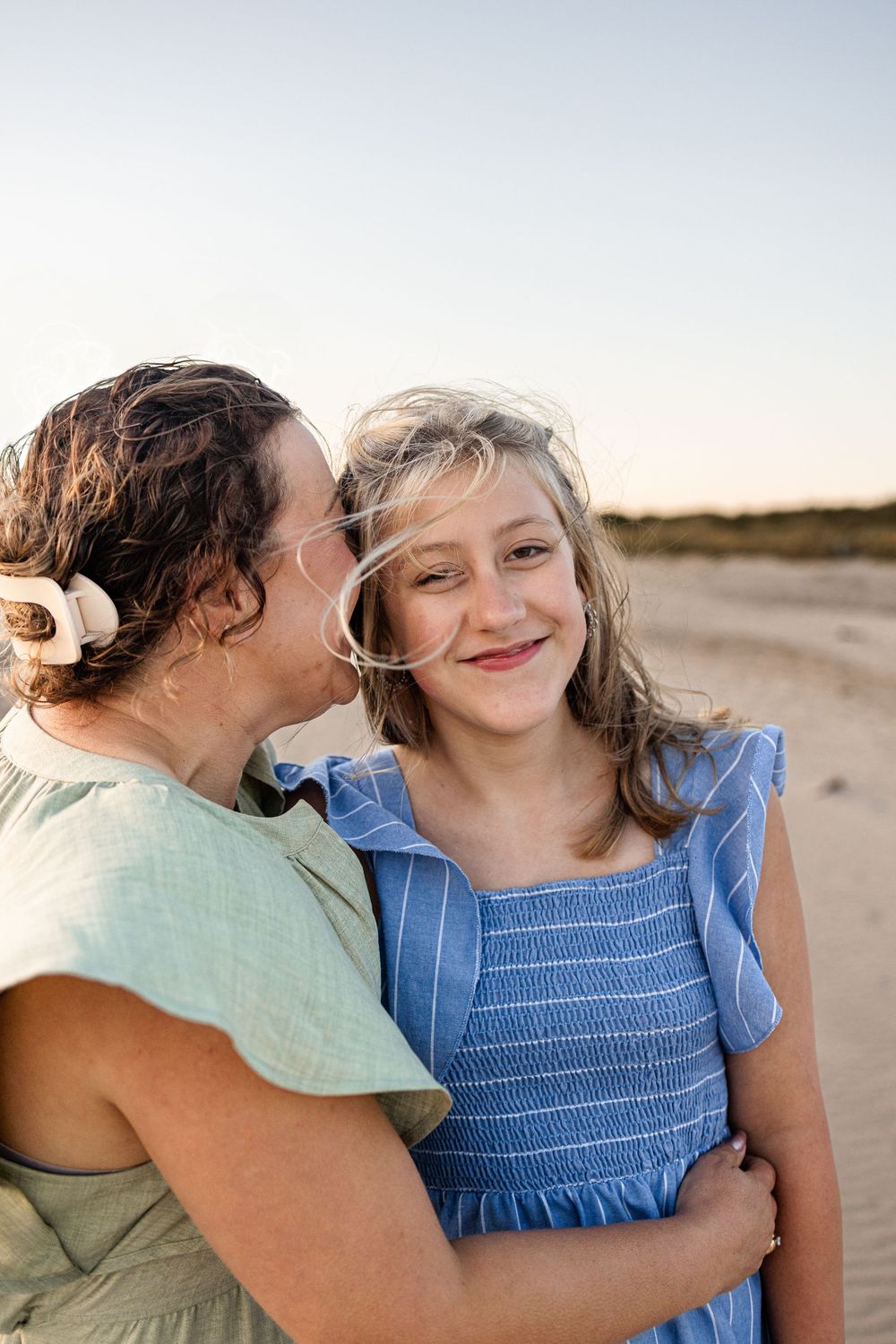 A parent and child share a tender moment on a sunny beach, both wearing blue and green casual clothing.