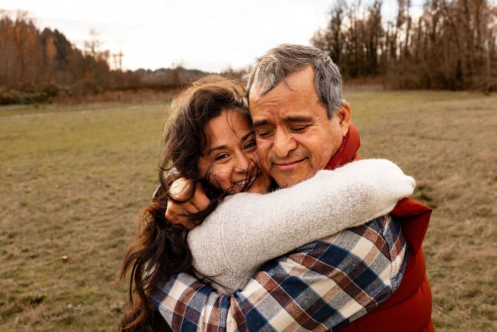 A woman and older man share a warm embrace outdoors in a field, smiling joyfully together.