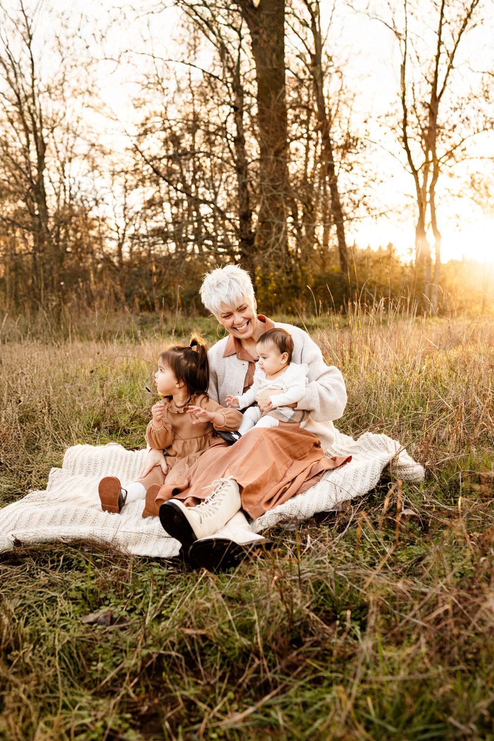 Woman with white hair sits on blanket in golden field at sunset, holding baby while young girl leans against her.