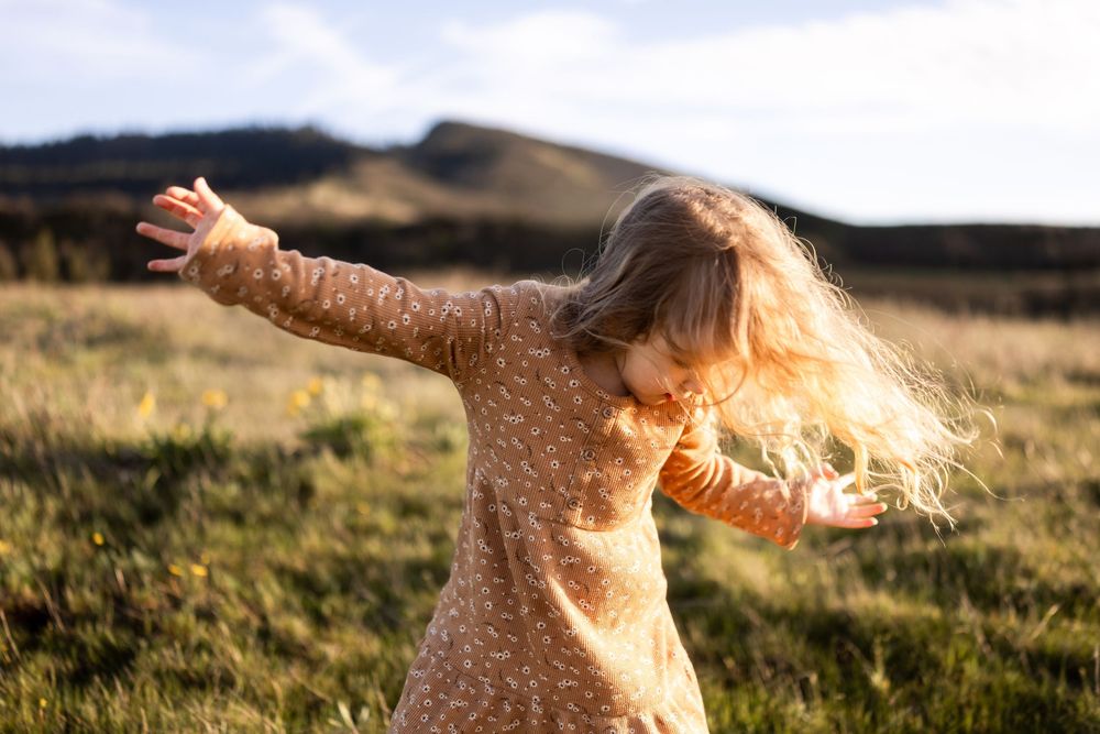 Young girl in floral dress twirling in a sunny meadow with hills in the background.
