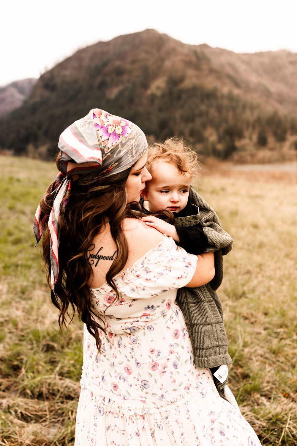 A bohemian mother in a floral dress and head scarf kisses her toddler in a scenic mountain meadow.