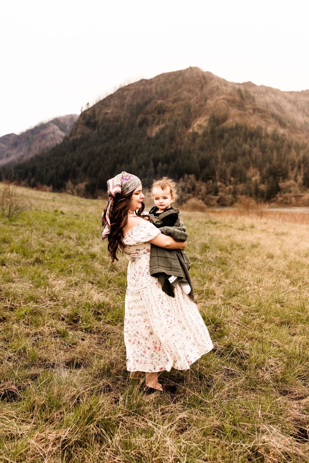 A mother in a floral dress holds her toddler in a green jacket in a mountain meadow.