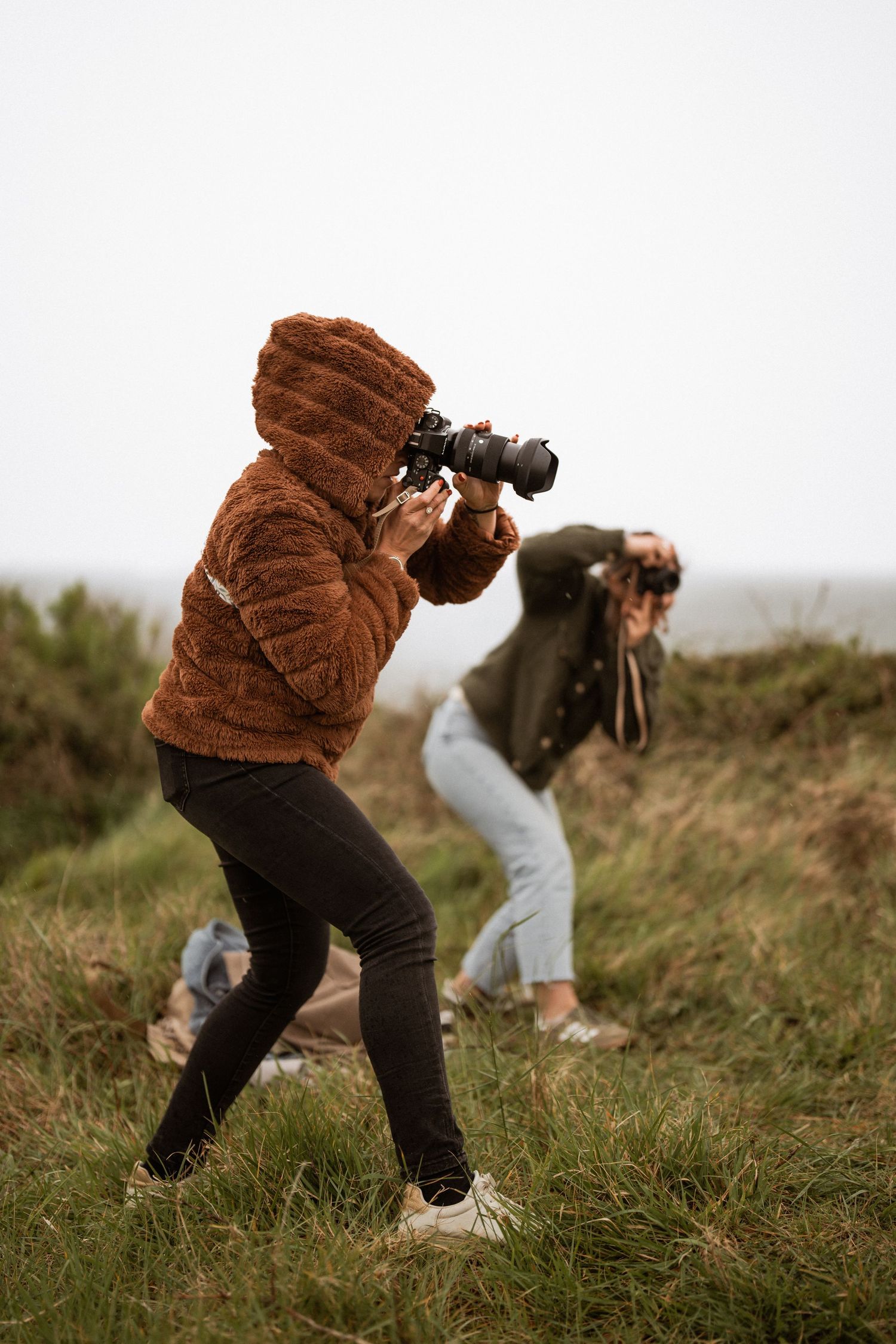 Photographer in brown jacket crouches in grassy field while taking pictures on an overcast day.