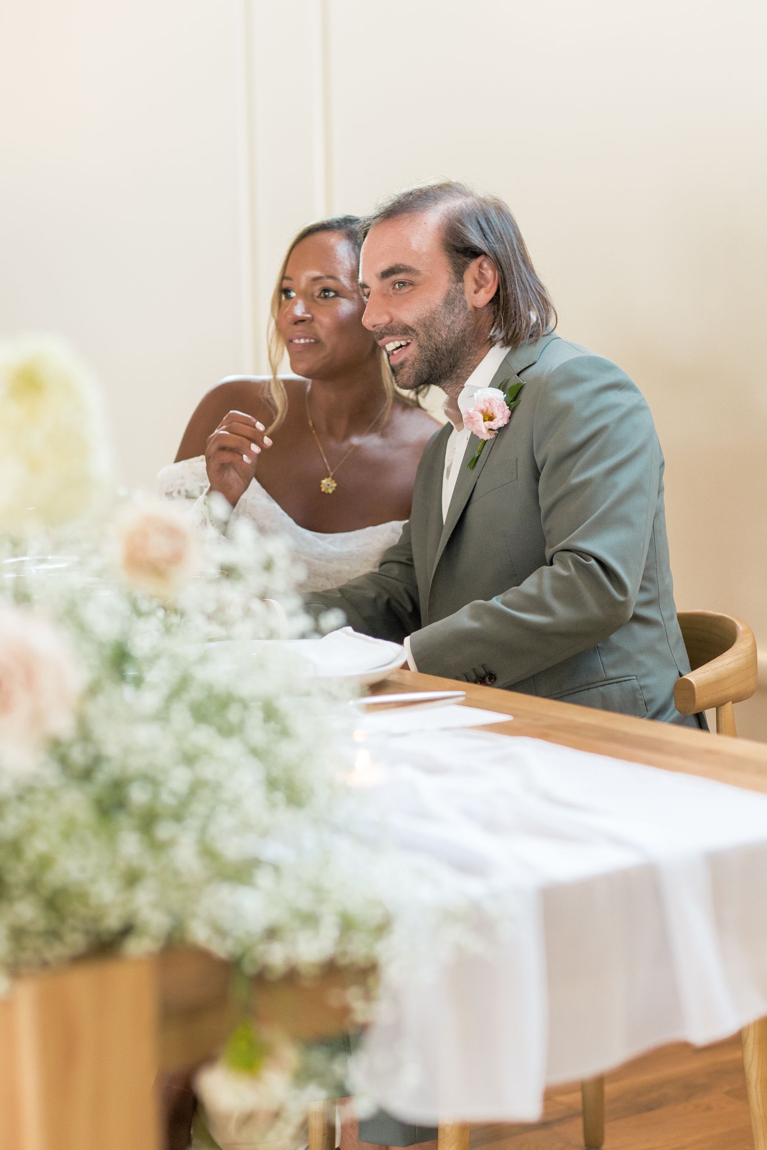 A couple seated at a table share an intimate moment, with white flowers in the foreground.