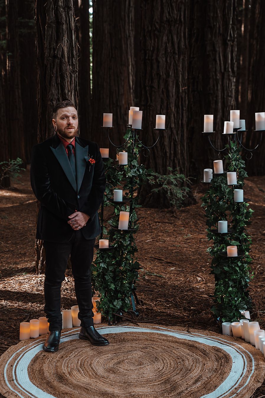 Person in dark formal attire stands in a forest setting with decorative candles and greenery at a wedding ceremony.