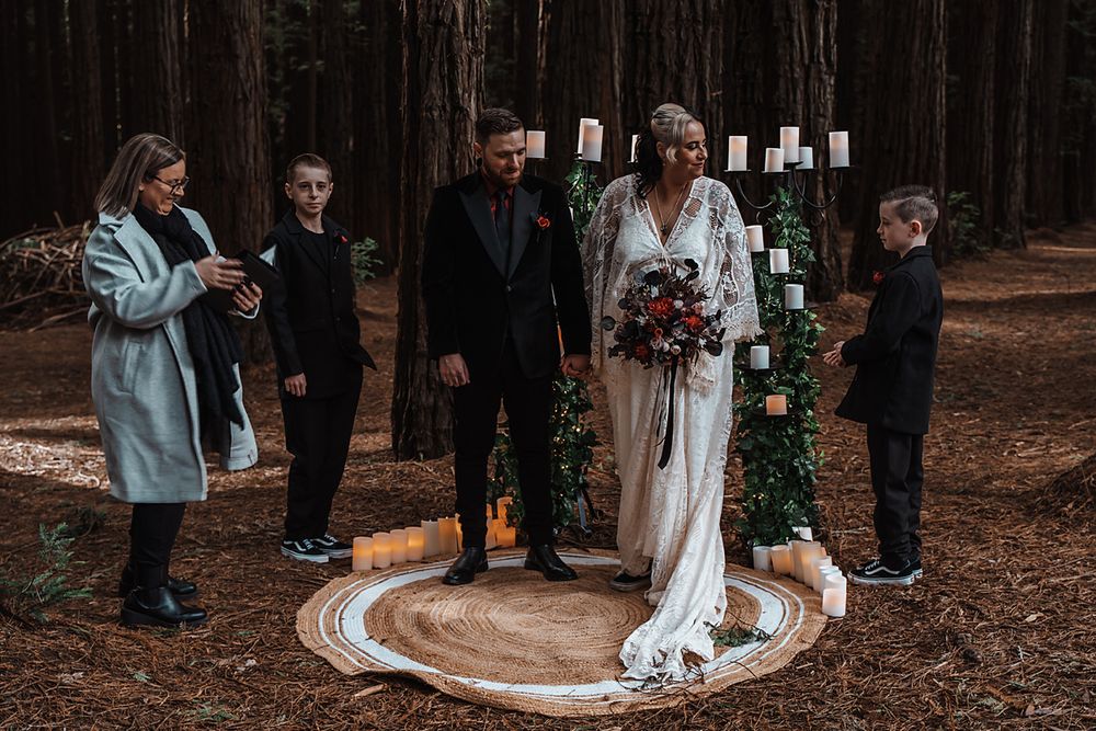 Wedding ceremony guests gathered in a forest setting with white candles and decorative greenery.